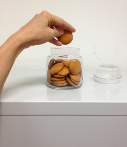 A woman's hand is suspended over a clear glass cookie jar. The jar is full of Vanilla Wafers, a small, disc shaped light brown cookie. The hand is holding a cookie (has just pulled one out of the jar). 